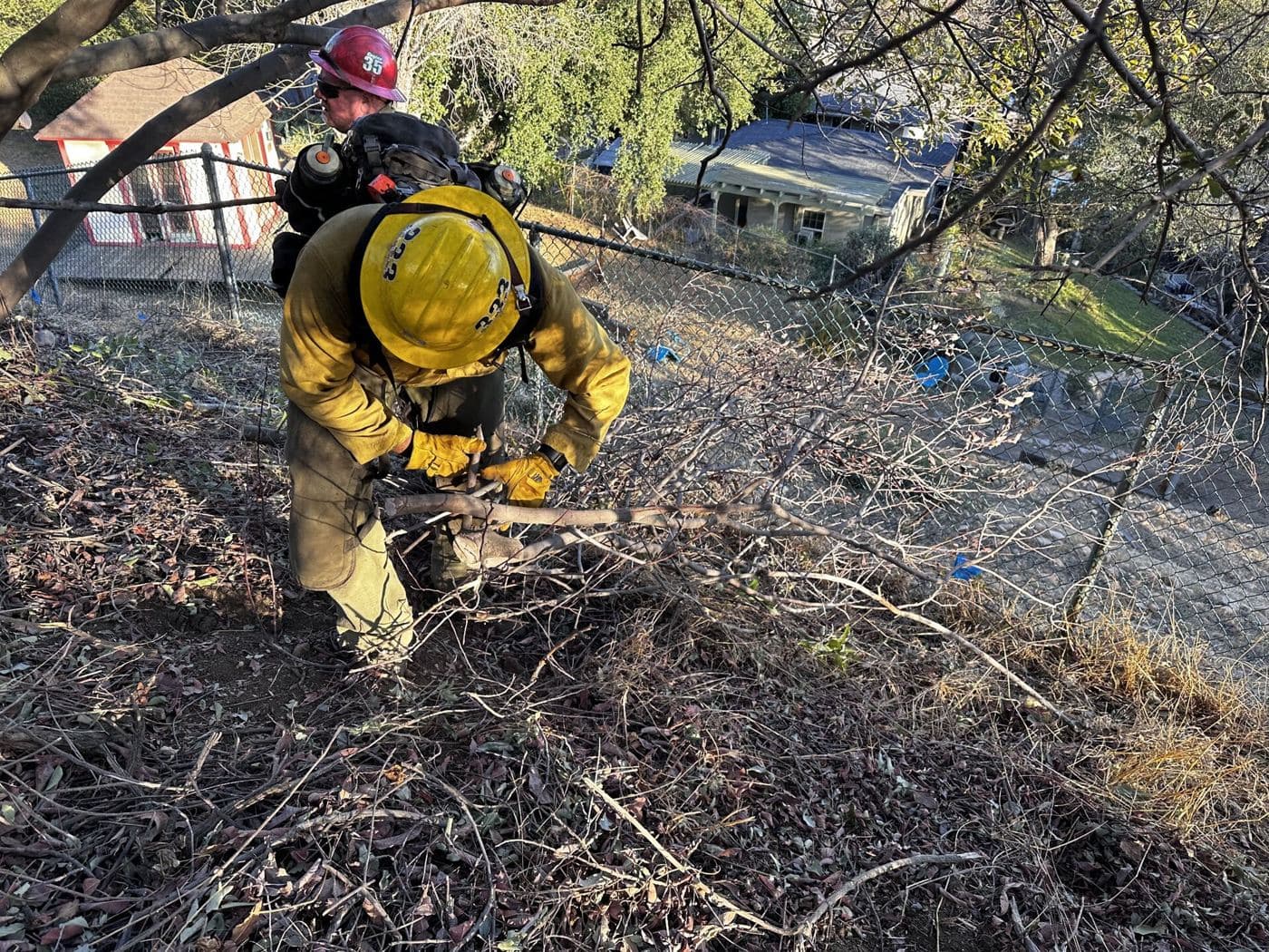 Firefighters cutting and clearing dry brush on a slope below a house.